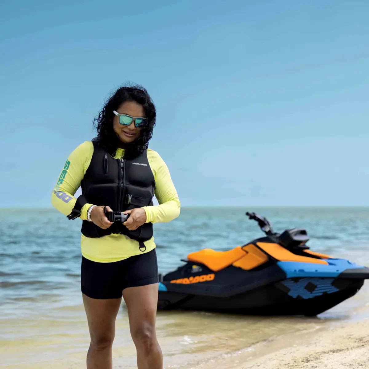 Sea-Doo Wave Life Jacket/PFD in black worn by a woman on a beach with a colorful jet ski in the background
