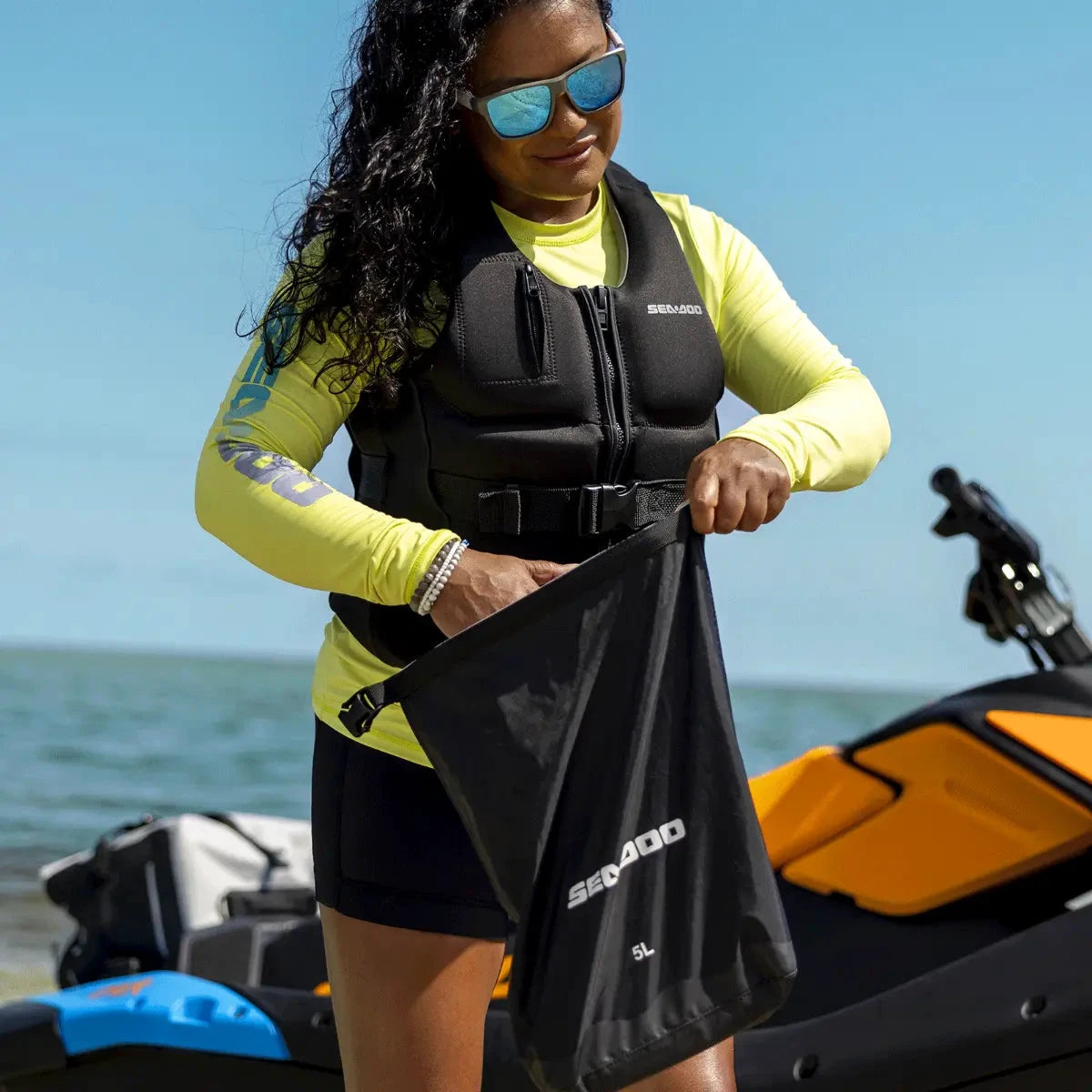 Sea-Doo 5L watertight roll-top bag, black with Sea-Doo logo, held near a jet ski.